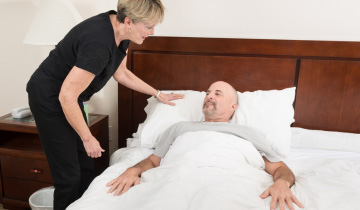 Eine Frau steht am Bett ihres pflegebedürftigen Mannes und kümmert sich um ihn