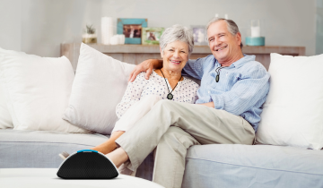 Zwei ältere Personen sitzen lächelnd auf einem Sofa und betrachten einen im Vordergrund stehenden Hausnotruf. Sie tragen einen Notfallknopf um den Hals.
